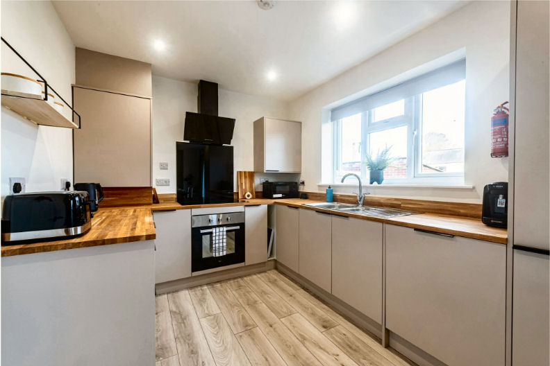 Modern kitchen with light wood flooring, white cabinets, wooden countertops, built-in oven, black appliances, large window, and a sink under the window.
