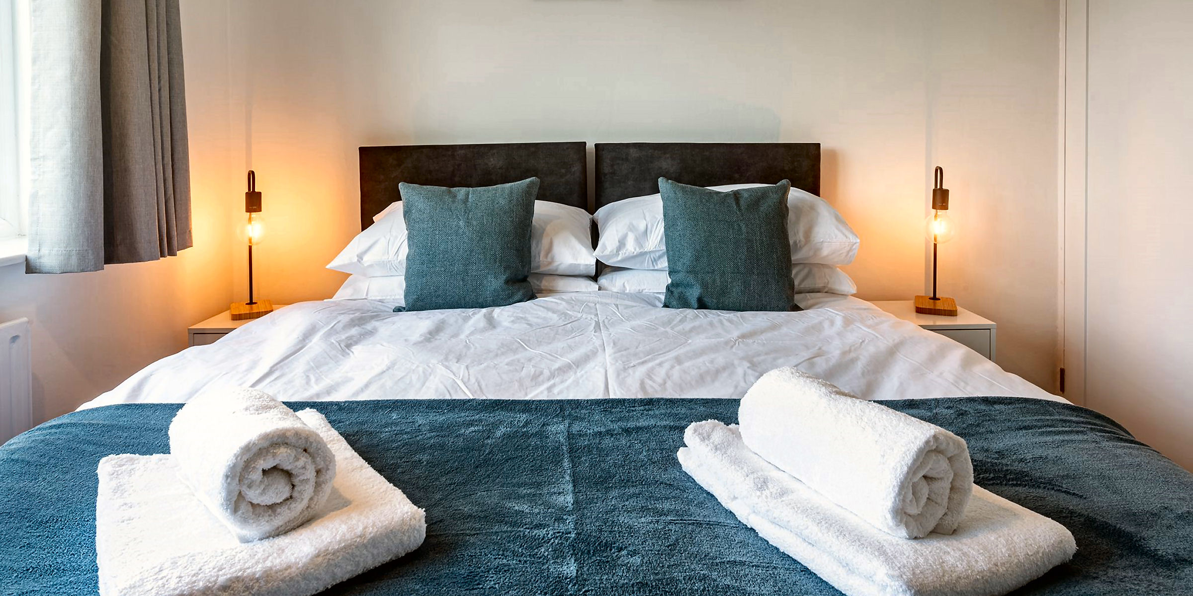 A neatly made bed with a dark headboard, white and teal bedding, two teal pillows, and two rolled white towels on a teal blanket. Warm-lit bedside lamps flank the bed.